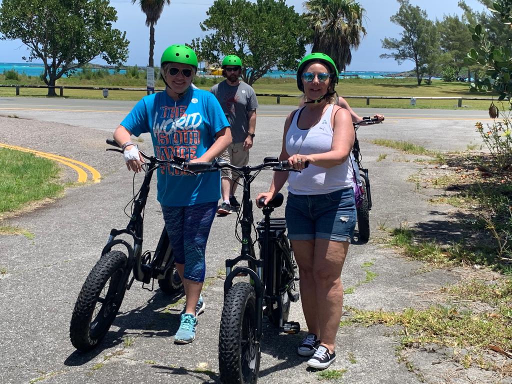 Two women wearing green helmets standing with electric bikes on a sunny road near the ocean.